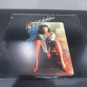 May include: A black vinyl record album cover with the title "Flashdance" in white text. The cover features a woman wearing a light blue sweater and red shoes, sitting on a couch.