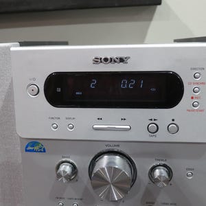 May include: Silver Sony stereo system with a digital display showing "2 0.21". Features include volume, bass, and treble controls, along with function buttons and a CD synchro option. The system has a sleek, modern design.