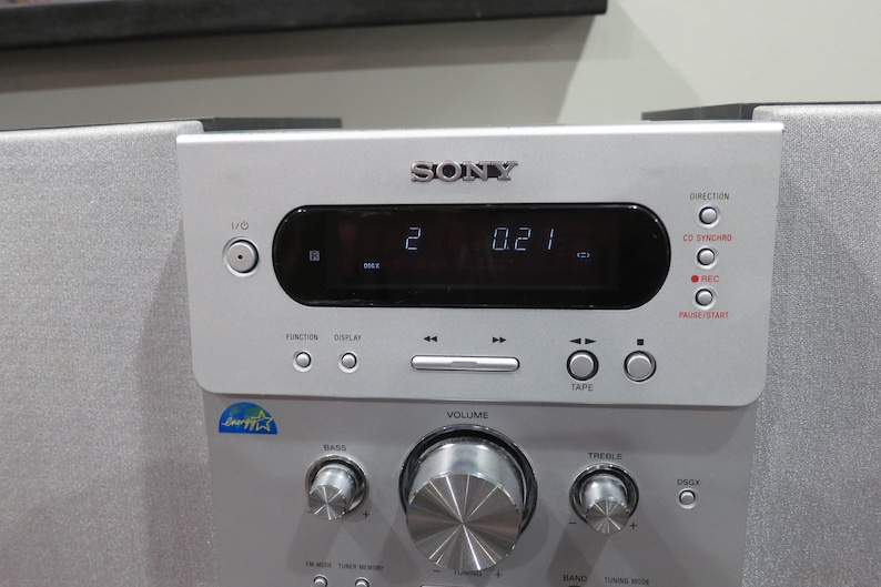 May include: Silver Sony stereo system with a digital display showing "2 0.21". Features include volume, bass, and treble controls, along with function buttons and a CD synchro option. The system has a sleek, modern design.