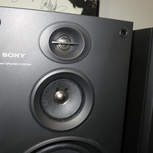 May include: Close-up of two black Sony 3-way speaker systems. The speakers have a matte finish and feature multiple circular speaker components. The Sony logo and "3 WAY SPEAKER SYSTEM" are printed on the front of the speaker.