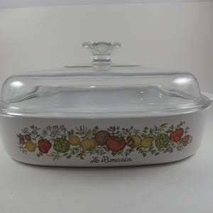 May include: A white rectangular ceramic casserole dish with a clear glass lid and a knob handle. The exterior features a colorful botanical print of various fruits and vegetables with the script text Le Romarin printed on the front side.