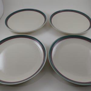May include: Four oval-shaped, off-white ceramic plates. Each plate has a decorative border of dark green and burgundy. The plates are arranged on a white surface, showcasing a simple, elegant design suitable for dining.