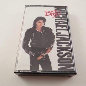 May include: A cassette tape featuring Michael Jackson's album "Bad." The cover art shows Jackson in a black jacket and pants. The album title is in red, and the artist's name is in large white letters.
