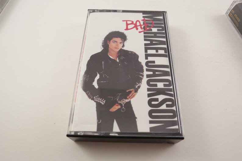 May include: A cassette tape featuring Michael Jackson's album "Bad." The cover art shows Jackson in a black jacket and pants. The album title is in red, and the artist's name is in large white letters.