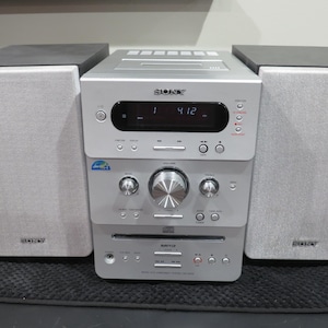 May include: A silver Sony stereo system with two gray speakers and a central unit. The central unit has a CD player, display, and various control buttons. The speakers have black tops and the system sits on a black mat.