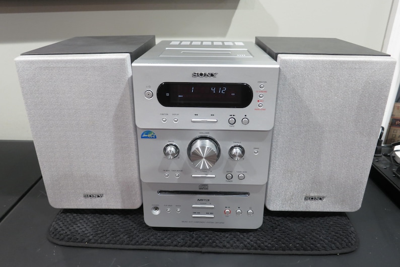 May include: A silver Sony stereo system with two gray speakers and a central unit. The central unit has a CD player, display, and various control buttons. The speakers have black tops and the system sits on a black mat.