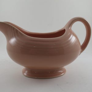 May include: A blush-colored ceramic gravy boat with a rounded body, a pouring spout, and a curved handle. The gravy boat has a pedestal base and a smooth, glossy finish. The design features concentric rings around the body.