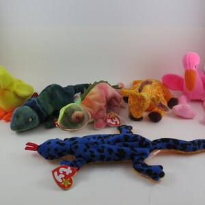 May include: A collection of colorful plush toys, including a yellow duck with an orange beak and feet, several lizards in shades of green, pink, and blue, a giraffe, and a pink flamingo. Each toy has a tag.