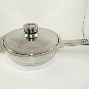 May include: A stainless steel saucepan with a matching lid and a long handle. The lid features a central knob with a circular design. The handle has a series of small, raised black dots near the base. The pan has a polished, reflective finish.