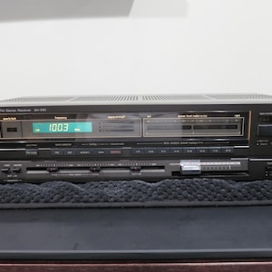 May include: A black Technics SA-250 AM/FM stereo receiver featuring a digital frequency display showing 100.3, a signal strength meter, power level meters, a headphone jack, and a horizontal volume slider control with various push-button input selectors.