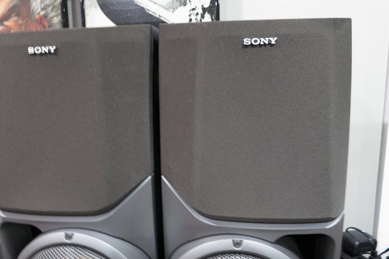 May include: Two gray Sony speakers with a silver Sony logo on the top. The speakers have a rectangular shape with a textured surface and a silver base. The bottom of the speakers have a circular speaker grill.