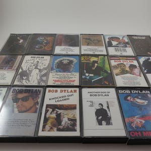 May include: A collection of 14 cassette tapes featuring Bob Dylan music. The tapes are arranged in three rows, with the titles of the albums visible on each tape. The titles include "Knocked Out Loaded", "Another Side of Bob Dylan", "Oh Mercy", "Planet Waves", "Blood on the Tracks", "Desire", "Street Legal", "Slow Train Coming", "Shot of Love", "Infidels", "Empire Burlesque", "Down in the Groove", "Under the Red Sky", and "Good as I Been to You".