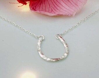 May include: A silver horseshoe necklace with a delicate chain. The horseshoe is textured and has a hammered finish. The necklace is displayed on a white background with pink flowers.