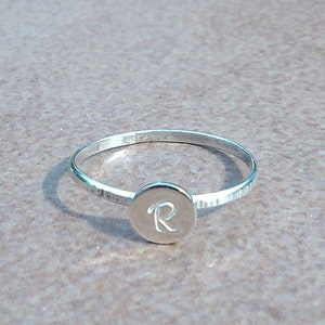 以下が含まれることがあります： 「R」の文字が刻まれた丸いディスクが付いた銀の指輪です。