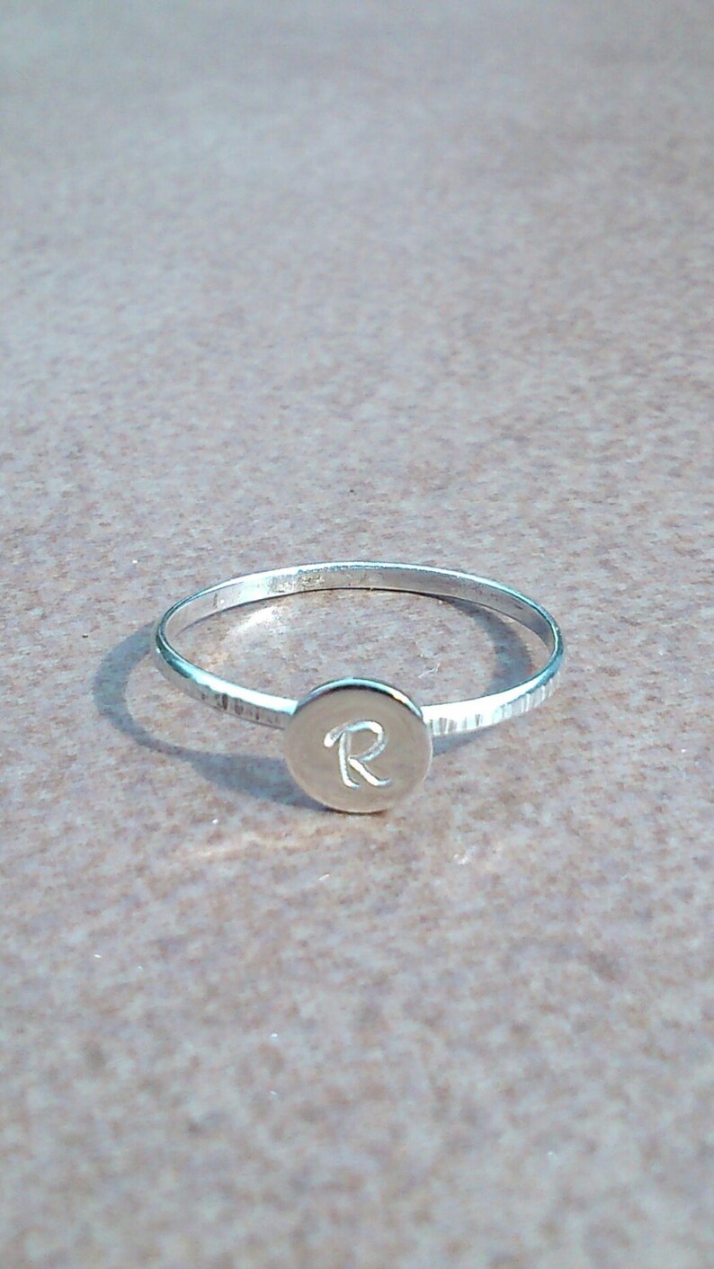 以下が含まれることがあります： 「R」の文字が刻まれた丸いディスクが付いた銀の指輪です。