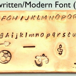 以下が含まれることがあります： 手書き/モダンフォント（2mm）と書かれた銅板。大文字と小文字の手書きフォントのアルファベット、矢印、十字架、サッカーボール、音符、波、トンボ、オームのシンボルなどが描かれている。