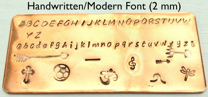 以下が含まれることがあります： 手書き/モダンフォント（2mm）と書かれた銅板。大文字と小文字の手書きフォントのアルファベット、矢印、十字架、サッカーボール、音符、波、トンボ、オームのシンボルなどが描かれている。