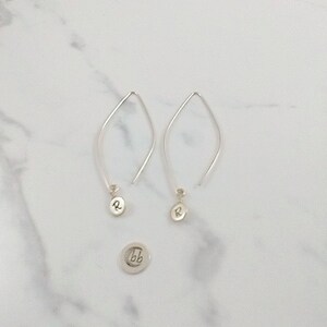 Puede incluir: Un par de pendientes de plata con un diseño delicado y curvo. Cada pendiente tiene un pequeño charm redondo con la letra "R" grabada.