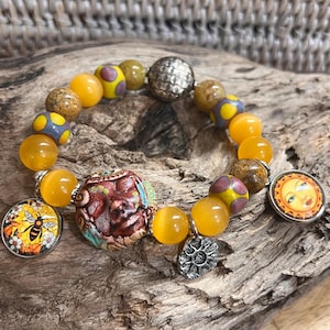 May include: A bracelet with a mix of yellow, brown, and silver beads. It includes a large silver bead, a charm with a bee, and a sun charm. The bracelet is displayed on a piece of wood. The beads are approximately 1 cm in diameter.