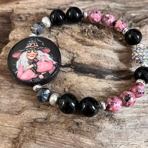 May include: A beaded bracelet featuring a round charm with a witch illustration. The bracelet includes black, pink, and silver beads. The witch charm is black with a pink and white witch illustration.