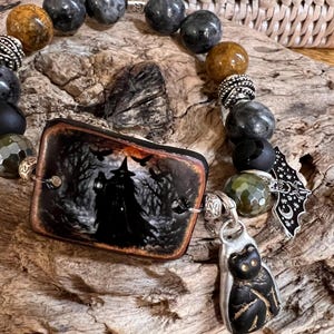 May include: A beaded bracelet featuring a rectangular focal piece with a witch and raven illustration, interspersed with grey, brown, and black beads, silver-toned spacers, a black cat charm, and a silver-toned bat charm with moon and star details.