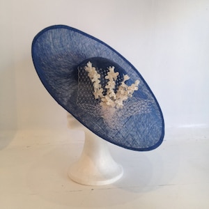 May include: A navy blue hat with a wide brim and a smaller, rounded crown. The hat is adorned with a cluster of white floral embellishments and a delicate veil. The hat is displayed on a white mannequin head.