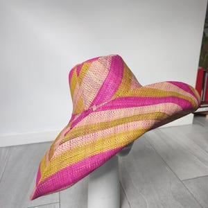 May include: A wide-brimmed straw hat featuring a woven pattern of alternating diagonal stripes in vibrant hot pink, mustard yellow, and natural beige. The hat has a structured crown and a broad, slightly undulating brim with a textured finish.