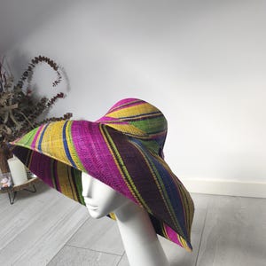 May include: A wide-brimmed woven hat featuring a vibrant striped pattern in shades of magenta, yellow, green, and dark blue. The hat is displayed on a white mannequin head, showcasing the textured material and the colourful, multi-directional stripes.