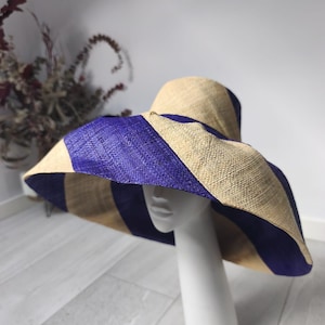 Pode incluir: Um chapéu de palha de aba larga com listras diagonais alternadas em bege natural e roxo profundo. O chapéu é exibido em uma cabeça de manequim branca, apresentando uma textura trançada em toda a copa e na aba larga e inclinada.