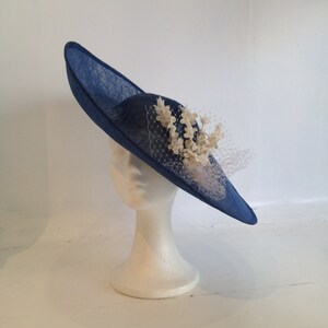 May include: A navy blue hat with a wide brim, displayed on a white mannequin head. The hat features a decorative arrangement of cream-colored flowers and a delicate veil. The hat's brim is approximately 15 inches in diameter.