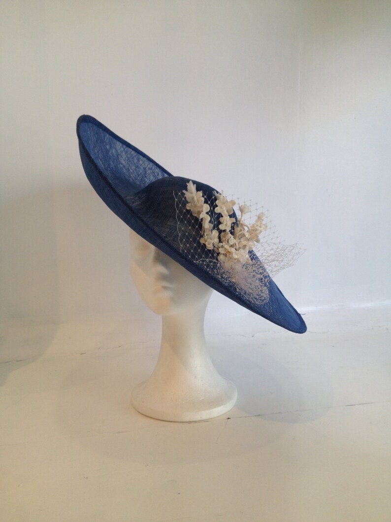 May include: A navy blue hat with a wide brim, displayed on a white mannequin head. The hat features a decorative arrangement of cream-colored flowers and a delicate veil. The hat's brim is approximately 15 inches in diameter.