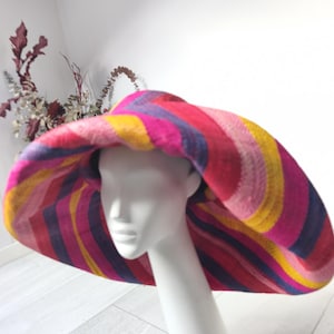 May include: A wide-brimmed sun hat featuring a vibrant, multi-coloured striped pattern in shades of hot pink, deep purple, bright yellow, and light pink. The hat is constructed from a woven material with a flexible, drooping brim and a rounded crown.