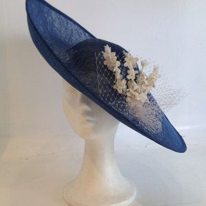 May include: A navy blue hat with a wide brim and a decorative cluster of off-white flowers and netting. The hat is displayed on a white mannequin head. The hat's brim is approximately 14 inches in diameter.