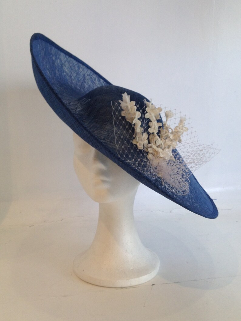 May include: A navy blue hat with a wide brim and a decorative cluster of off-white flowers and netting. The hat is displayed on a white mannequin head. The hat's brim is approximately 14 inches in diameter.