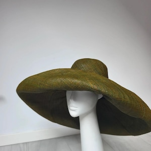 Peut inclure: Un chapeau à larges bords de couleur vert olive foncé avec une texture en paille tressée. Le chapeau présente une calotte arrondie et un grand bord évasé qui s'étend vers l'extérieur, présenté sur une tête de mannequin blanche unie.