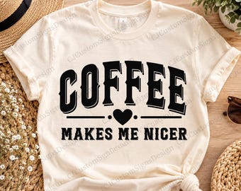May include: A cream-colored short-sleeve t-shirt featuring black text that reads COFFEE in a bold font, a small black heart icon, and the text MAKES ME NICER below it. The design is centered on the front of the garment.