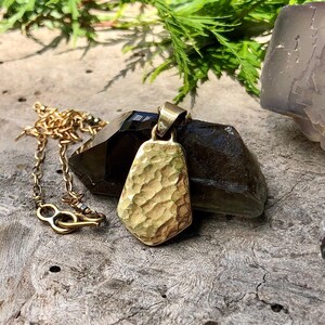 May include: A gold necklace with a hammered brass pendant. The pendant is a teardrop shape and has a textured surface. The necklace is on a wooden surface with a clear quartz crystal.