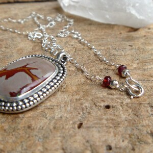May include: A silver necklace with a red and brown stone pendant. The pendant is set in a silver bezel and has a beaded edge. The chain is silver and has a lobster clasp.