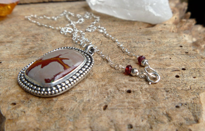 May include: A silver necklace with a red and brown stone pendant. The pendant is set in a silver bezel and has a beaded edge. The chain is silver and has a lobster clasp.