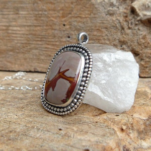 May include: A silver pendant with a brown and red agate stone set in a beaded bezel. The pendant is on a silver chain.