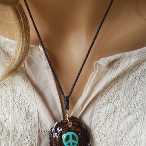Puede incluir: Un collar con un colgante de signo de paz. El colgante está hecho de resina marrón y tiene un signo de paz turquesa en el centro. El collar está hecho de cordón negro.