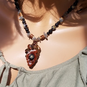 May include: A brown and black necklace with a carved wooden pendant featuring a flower design. The necklace is made with lava rock, wood, and glass beads.