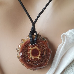 Può includere: Un ciondolo in legno marrone con un design a sole rosso e bianco, appeso a un cordoncino nero.