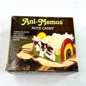 May include: A boxed Ani-Memos Note Caddy featuring a cow and rainbow design. The caddy holds a stack of notepaper and is decorated with a cow, rainbow, and sun. The box reads "COMPLETE WITH 200 SHEETS OF COLOR-COORDINATED PAPER."