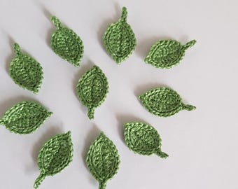 May include: A collection of ten small, crocheted green leaves arranged on a white surface. Each leaf has a detailed texture and a small stem, suggesting a handmade craft project or decorative element.