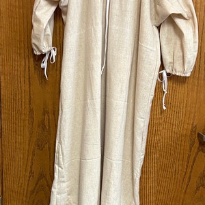 May include: A long, beige linen dress with three-quarter sleeves and a gathered neckline. The dress features white ties at the neckline and sleeve cuffs. It hangs from a clothes hanger and is displayed against a wooden door.