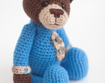 May include: A crocheted brown bear wearing a blue crocheted outfit with brown buttons. The bear is sitting with its legs crossed.