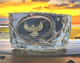 May include: A silver, copper, and gold bracelet with a blue stone and a gold winged figure in the center. The bracelet is decorated with swirls and dots.