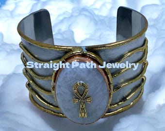 May include: A silver and gold cuff bracelet with a white oval stone featuring a gold ankh symbol. The bracelet has a copper accent and the text "Straight Path Jewelry" is visible.