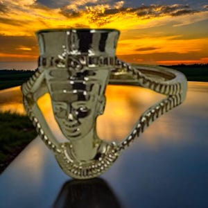 Peut inclure: Une bague en or avec un design détaillé de la tête d'un pharaon égyptien. La bague est placée sur un fond de coucher de soleil au-dessus d'une étendue d'eau.